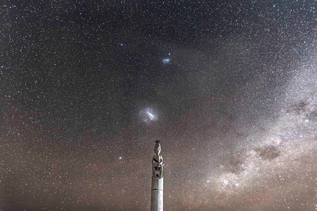 Star-filled night sky with a tall carved statue in the foreground and a bright galaxy nearby.