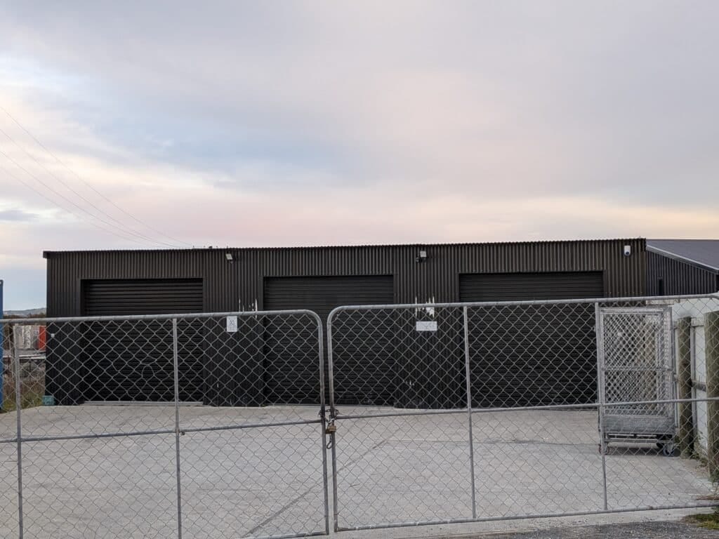 Industrial corrugated metal building behind chain-link fence with power lines and overcast sky.