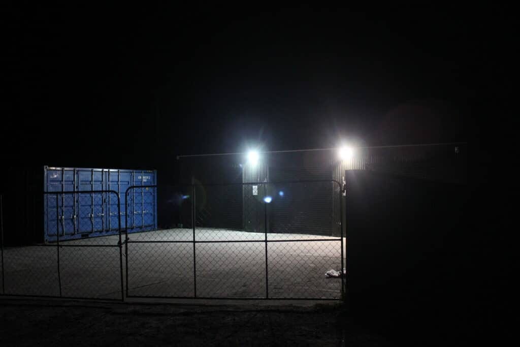 Nighttime industrial yard with blue shipping containers, corrugated building, and bright exterior lights.