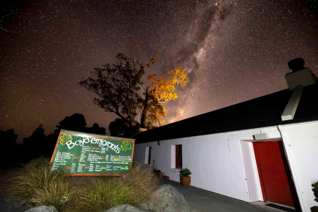 Milky Way and starry sky above a white building with red door, illuminated trees, and chalkboard sign.
