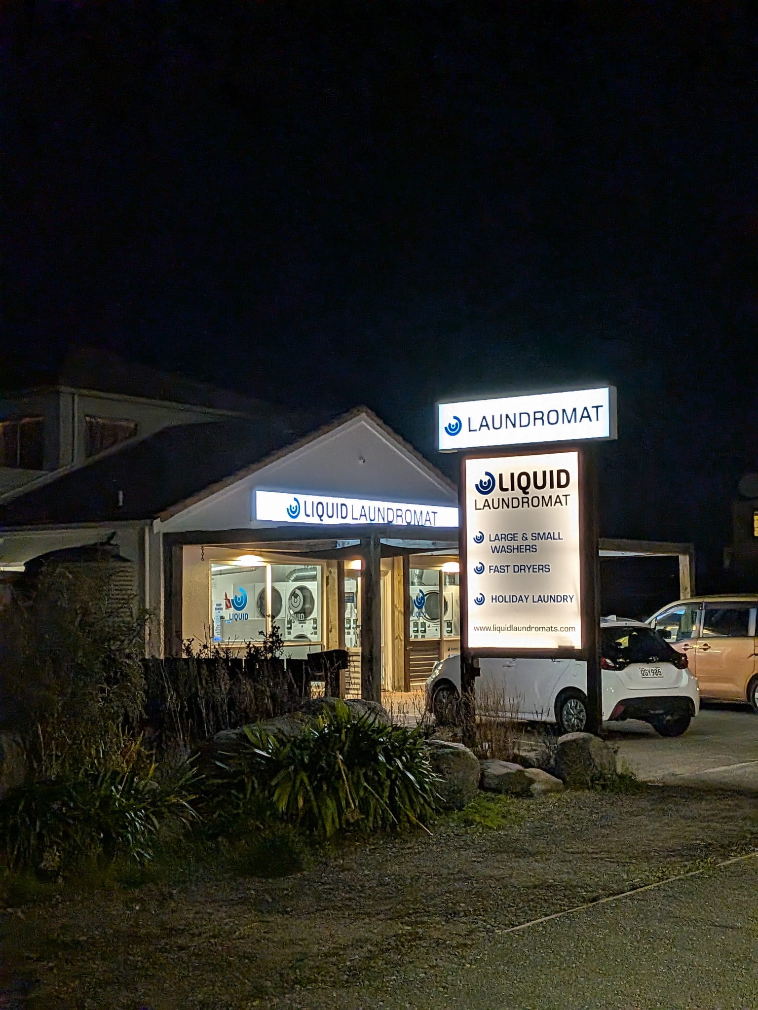 Nighttime view of a small laundromat with illuminated signs, a white car, and surrounding shrubs.