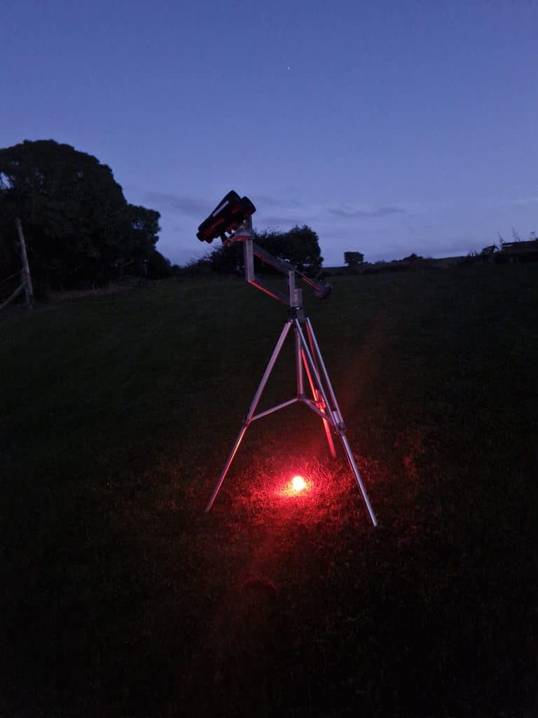 Telescope on a tripod with red illumination in a grassy field at dusk.