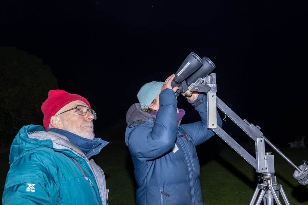 Two people outdoors at night using a large binoculars on a metal tripod, looking up.