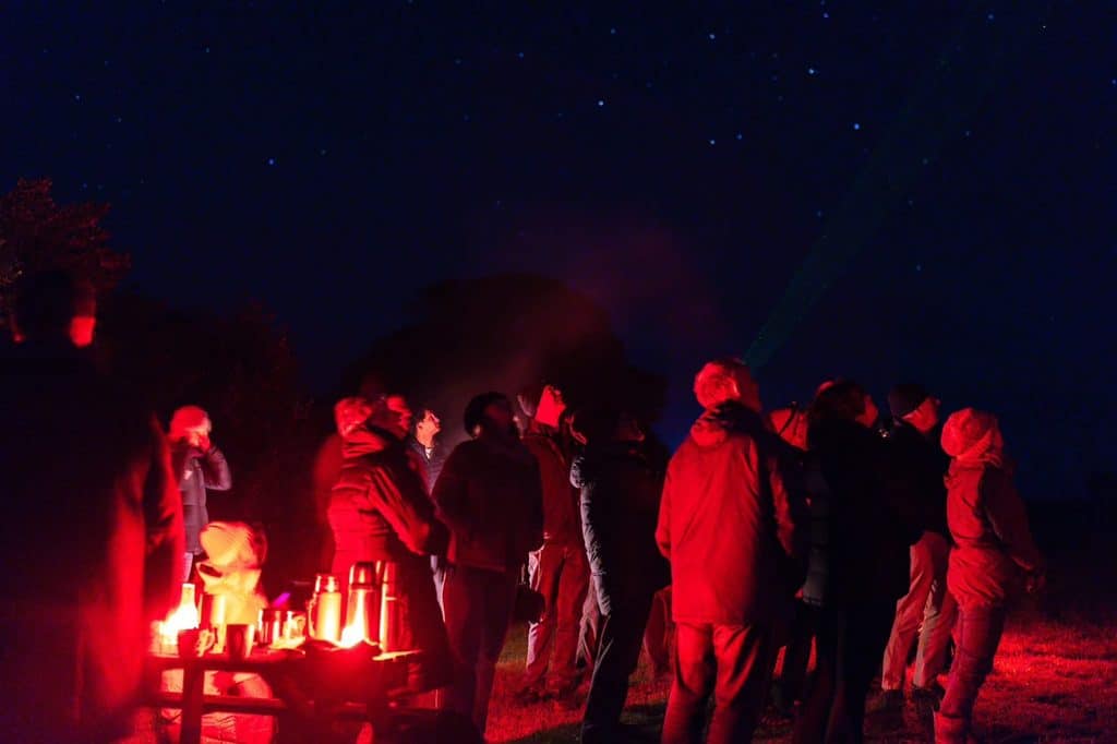 Group of people in warm jackets around a red-lit campfire at night, starry sky above.