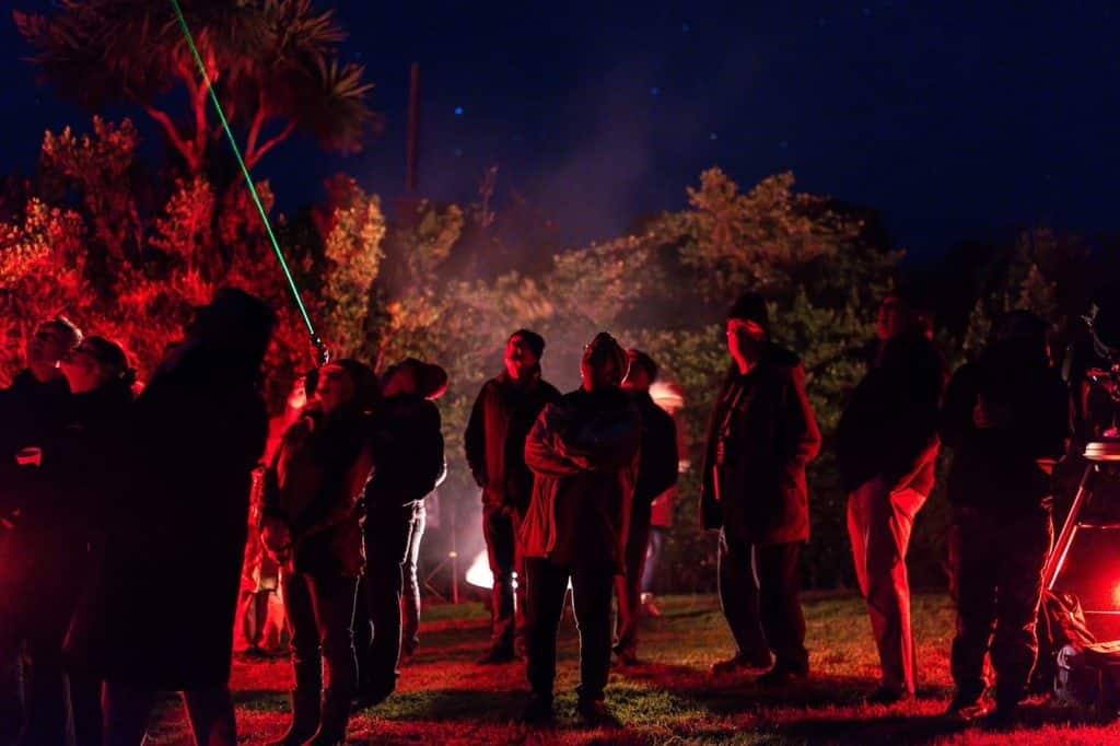 Group of people outdoors at night, red/green lights, standing on grass with trees, some looking up.