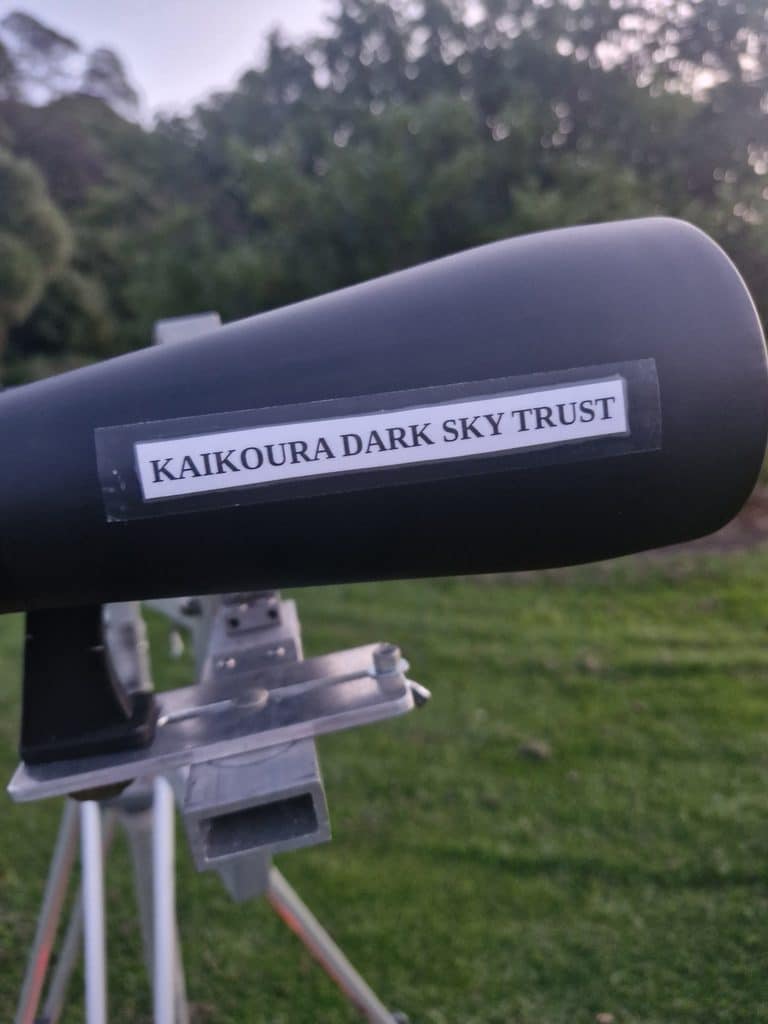 Telescope or viewing instrument on a metal stand labeled “Kaikoura Dark Sky Trust” outdoors.