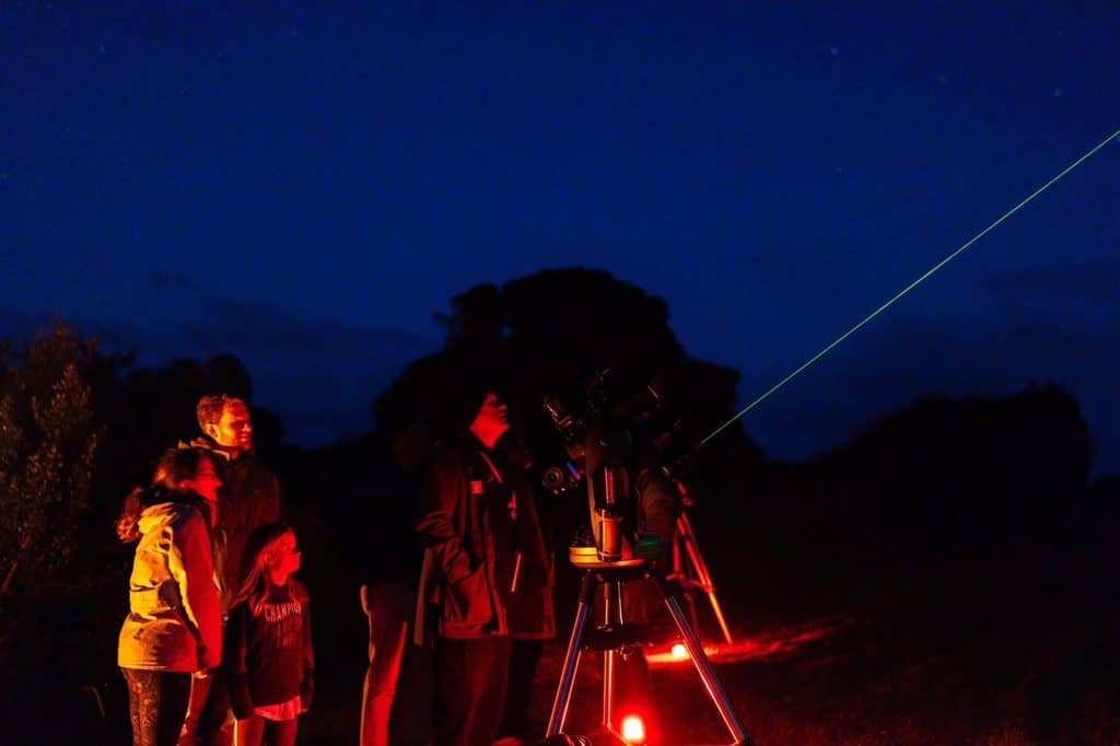 Group of people with a telescope at night, green laser line across dark sky.