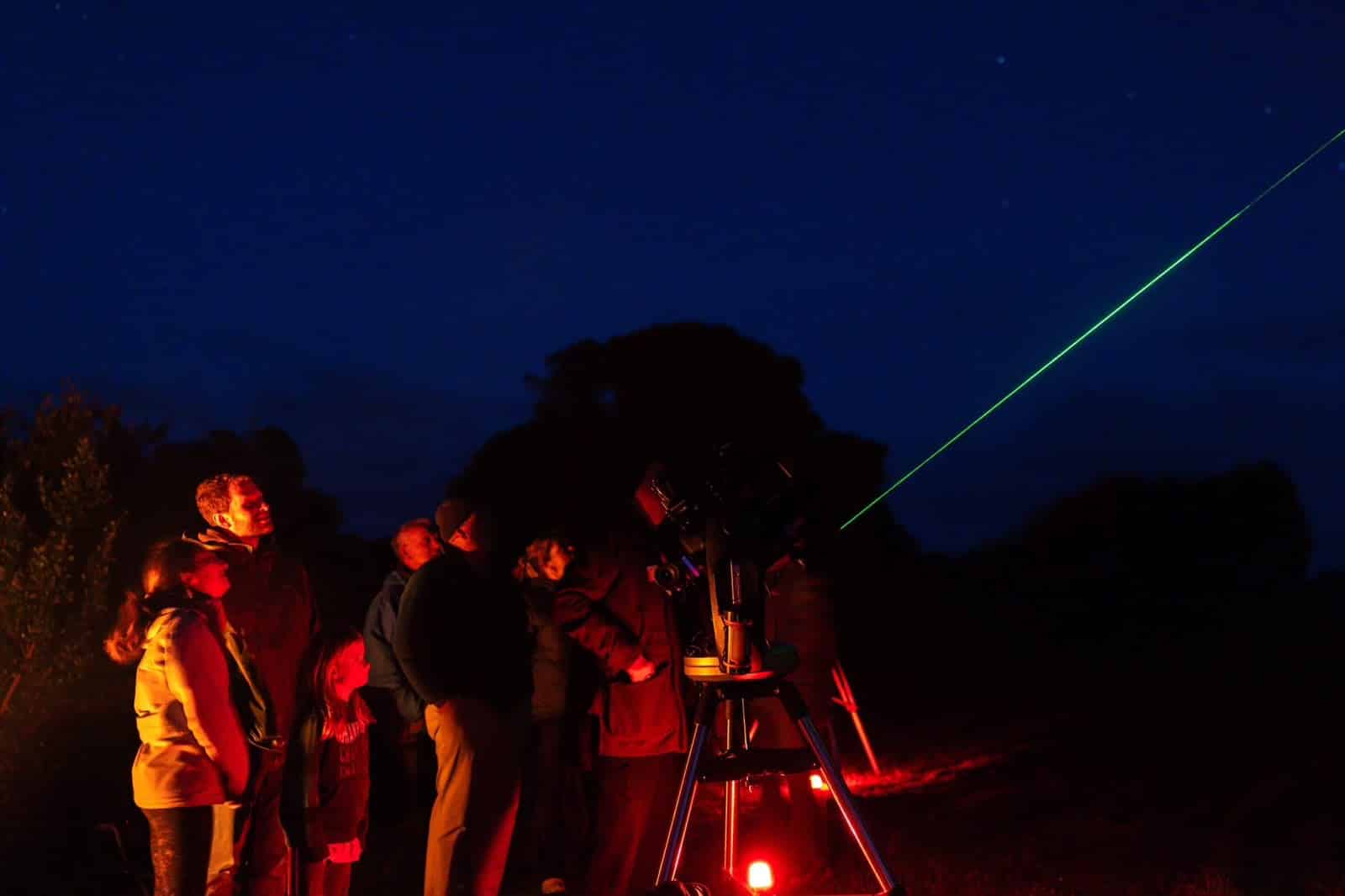 Group of people outdoors at night watching a green laser beam with a telescope on a tripod nearby.