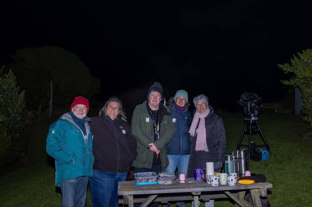 Group of five adults outdoors at night, with a telescope and table of drinks and snacks.