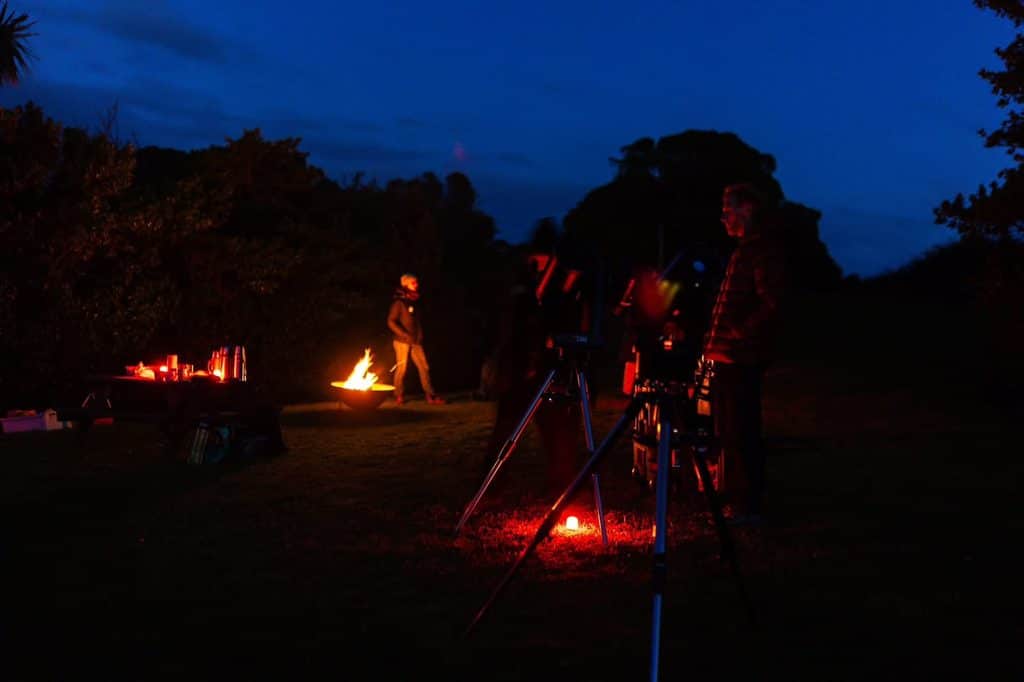 Night outdoor gathering around a campfire with people, equipment, and red glow.